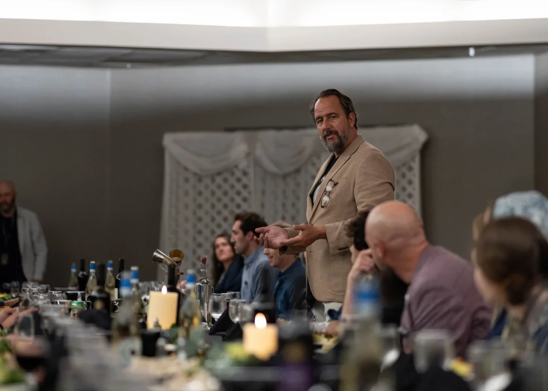 A man speaks with an open hand as people listen beside him at the table.
