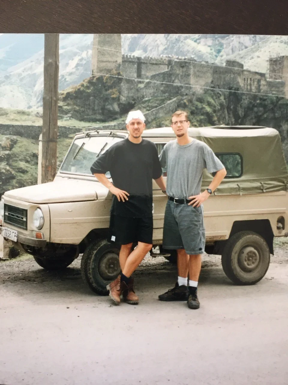 Two men stand beside a beige off-road vehicle, with stone fortifications and mountains behind them.