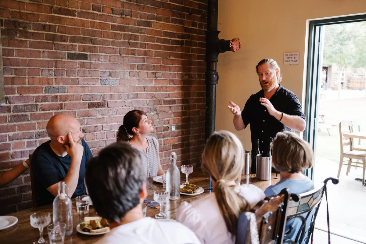 A standing man gestures as guests seated around a restaurant table listen.