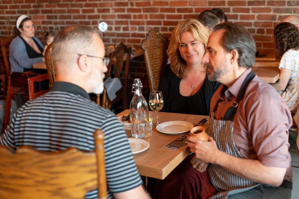 A man wearing an apron holds a small clay vessel while talking with two people at a restaurant table.