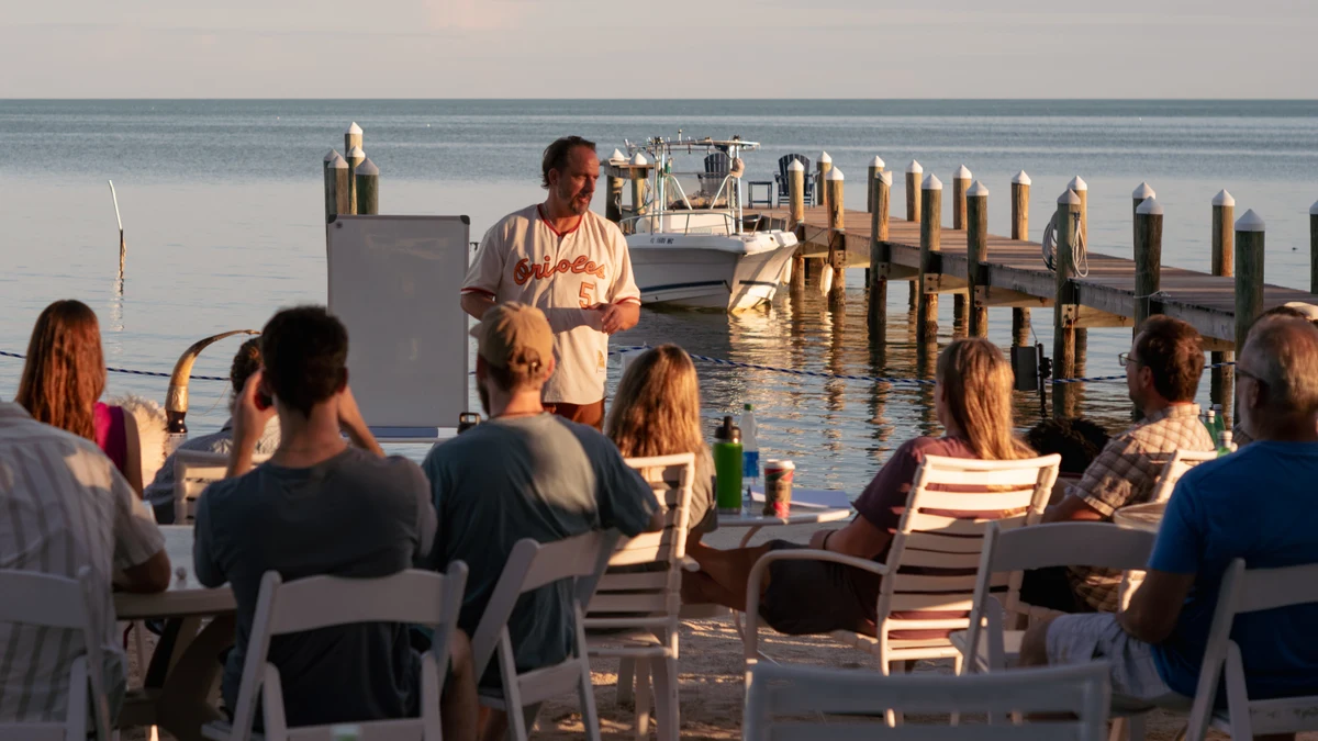 A man stands beside a whiteboard addressing a seated group beside the water, with a boat and dock behind him.