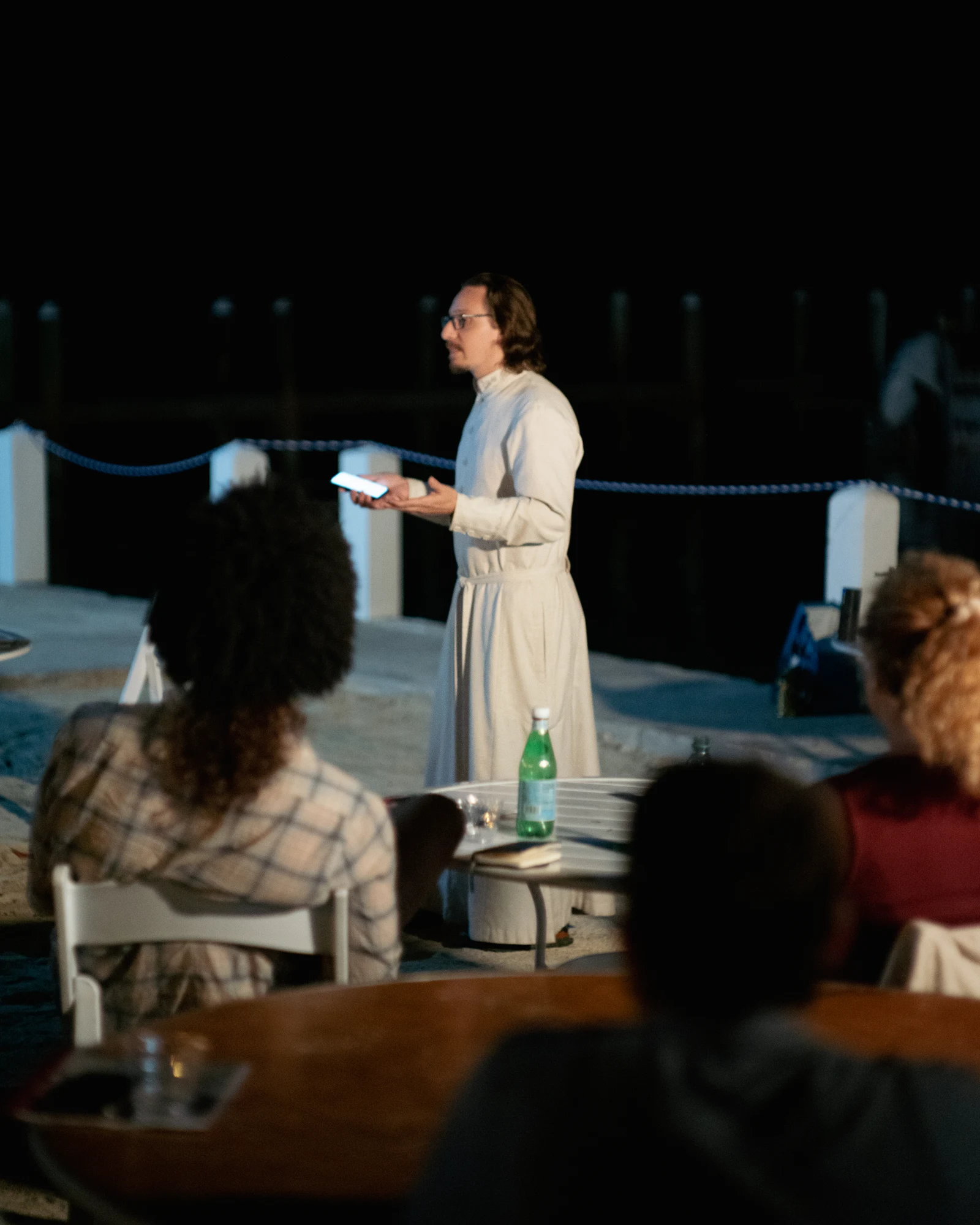 A presenter holds a phone and gestures while addressing seated listeners outdoors at night.