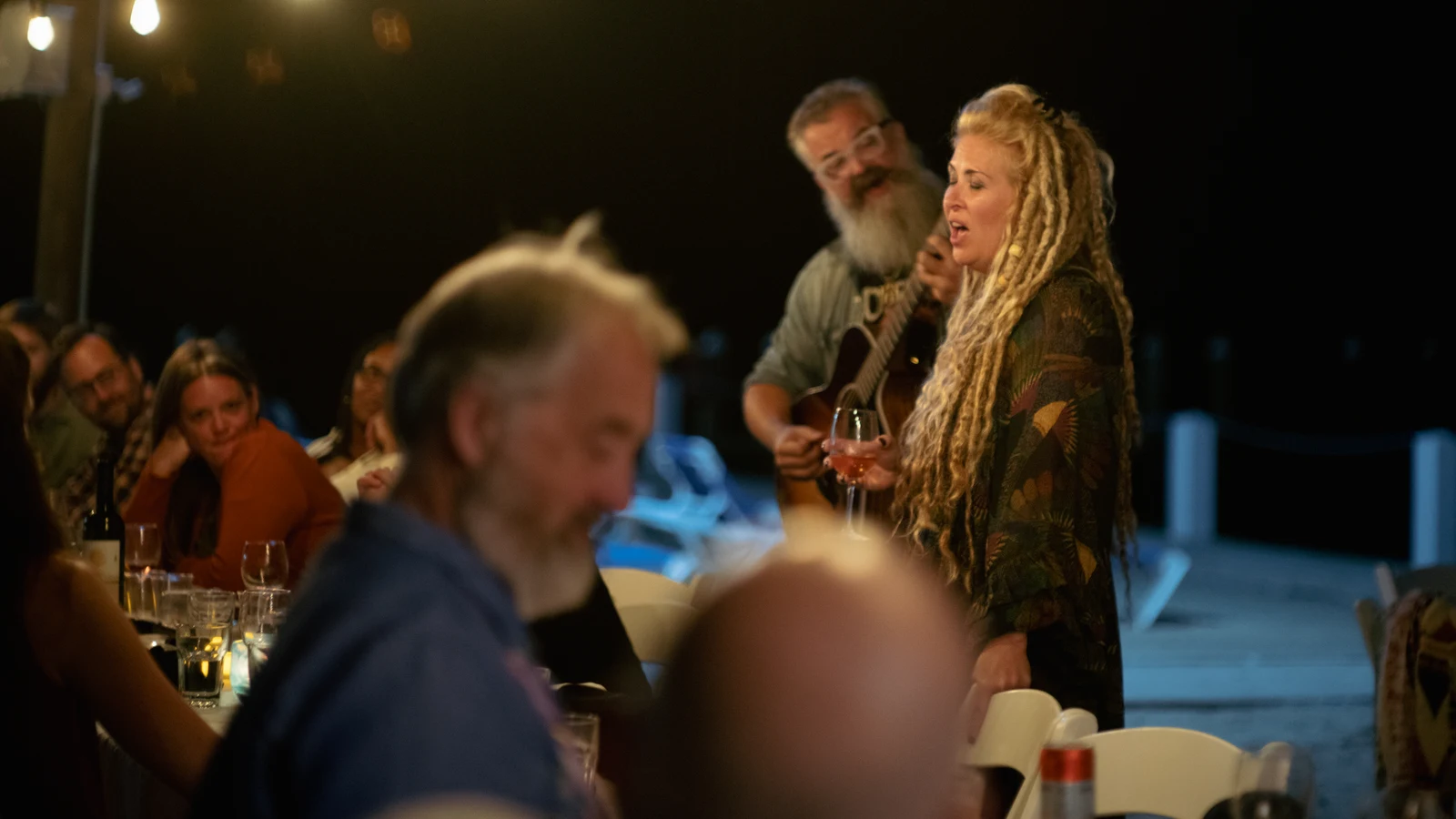 A woman sings beside a man playing an acoustic guitar as guests sit around a long dinner table.