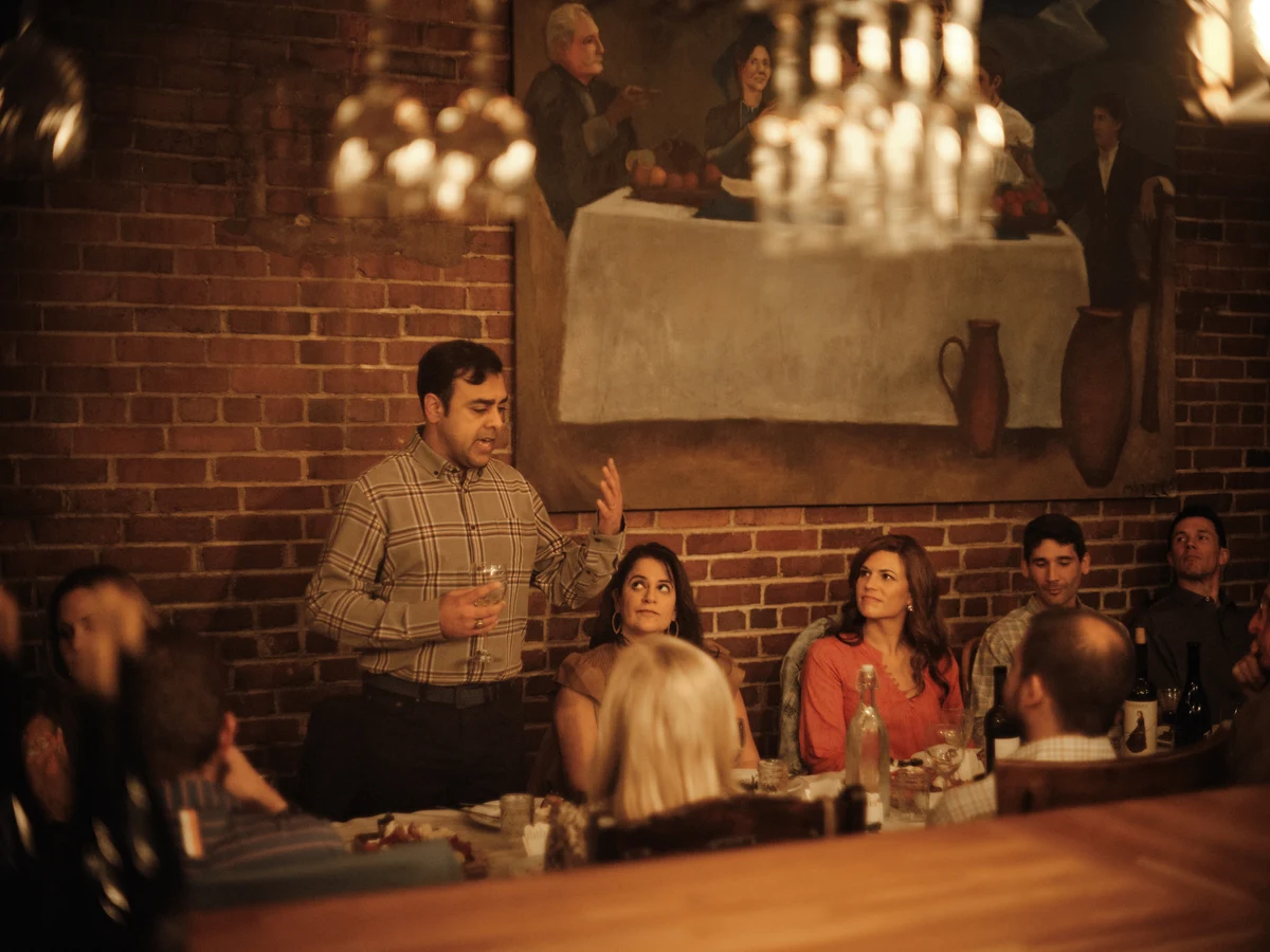 A standing guest holds a glass and speaks while people seated around a long table look toward him.
