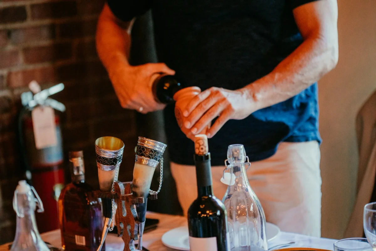 A person uses a corkscrew on a wine bottle beside glassware and drinking horns on a table.