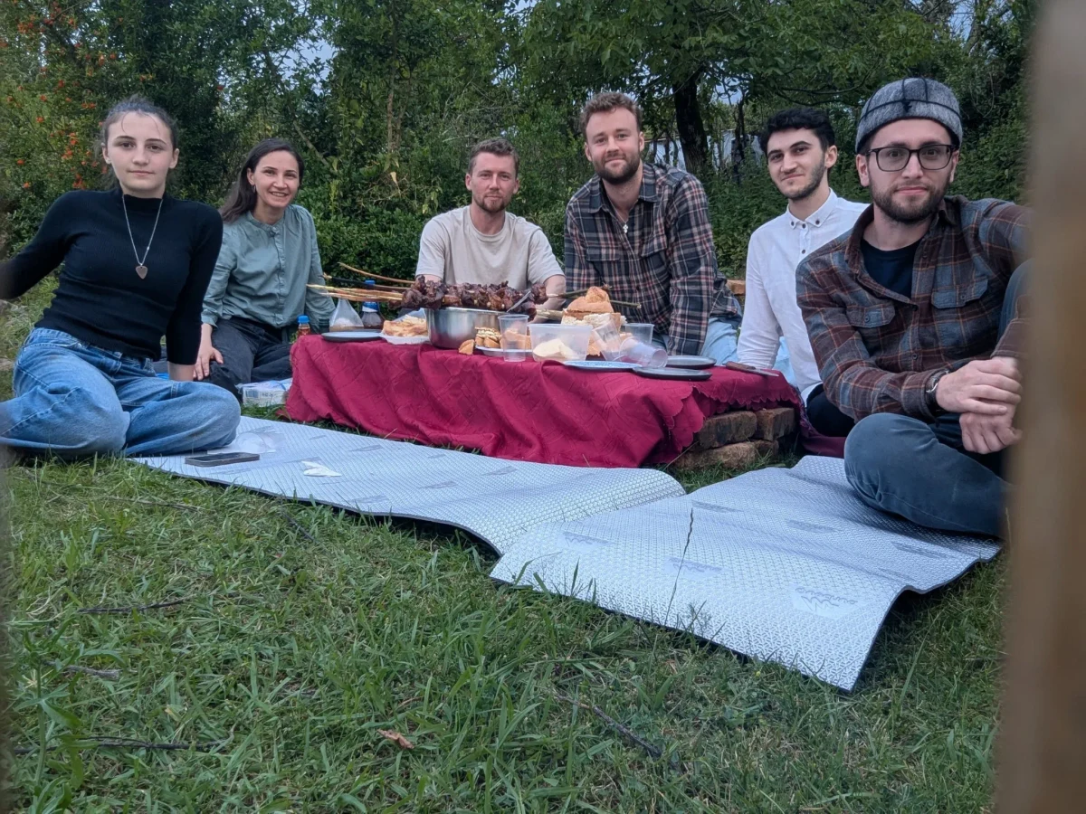 Six people sit outdoors around a low table covered in red cloth, with shared food beneath the trees.
