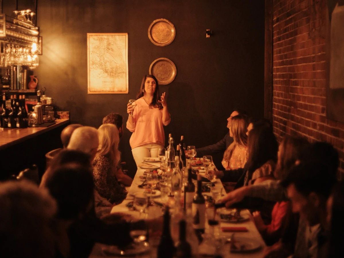 A woman holding a glass speaks and gestures while guests seated around the dinner table watch her.