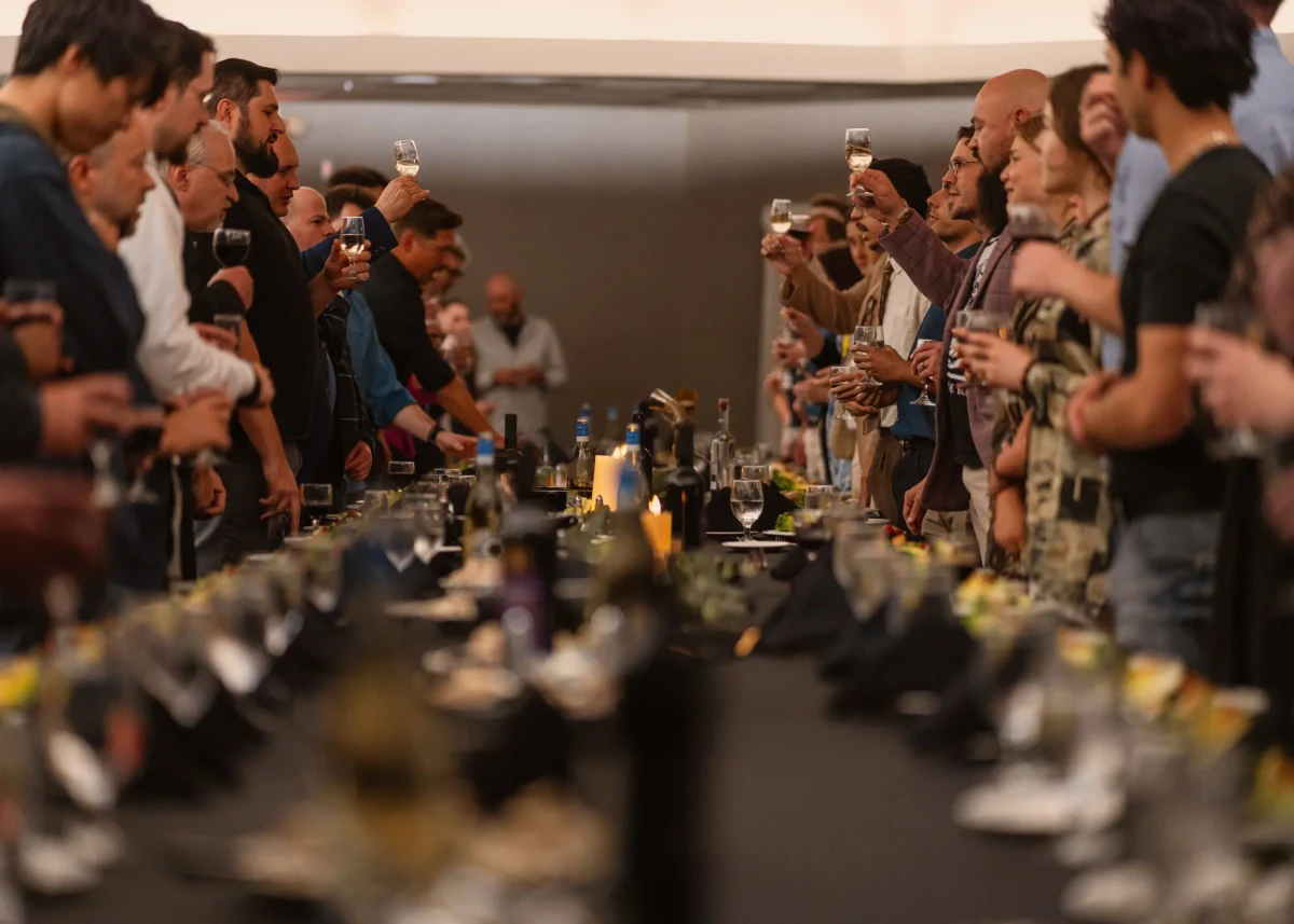 People standing on both sides of a long dinner table raise their glasses for a toast.