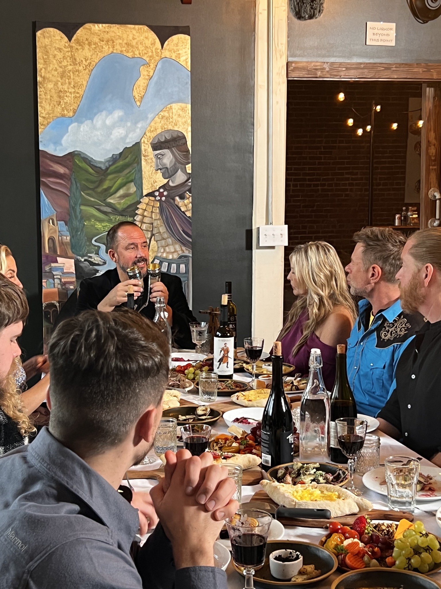 A man speaks while guests listen around a table set with food and drinks.